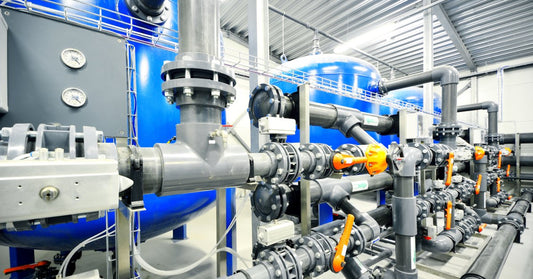 An industrial boiler room with stainless steel pipework and large blue boiler tanks. The pipework valves are orange.