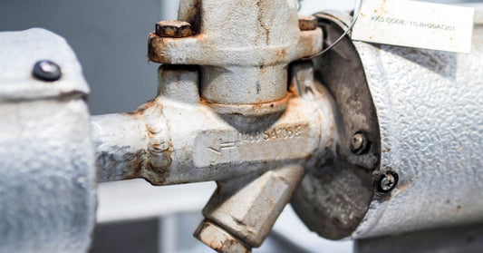 A close-up of a white, mildly rusty steam trap situated between two white pipe systems to trap condensate.