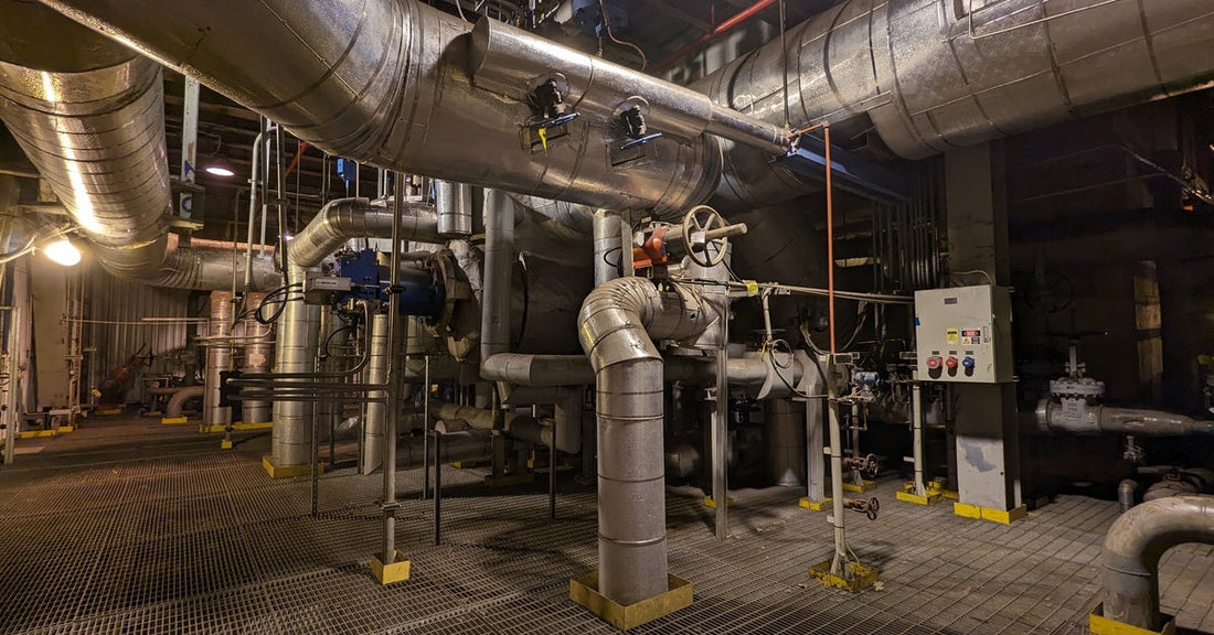 An industrial steam boiler room with stainless steel pipework and underground flood lighting. There are vents on the floor.